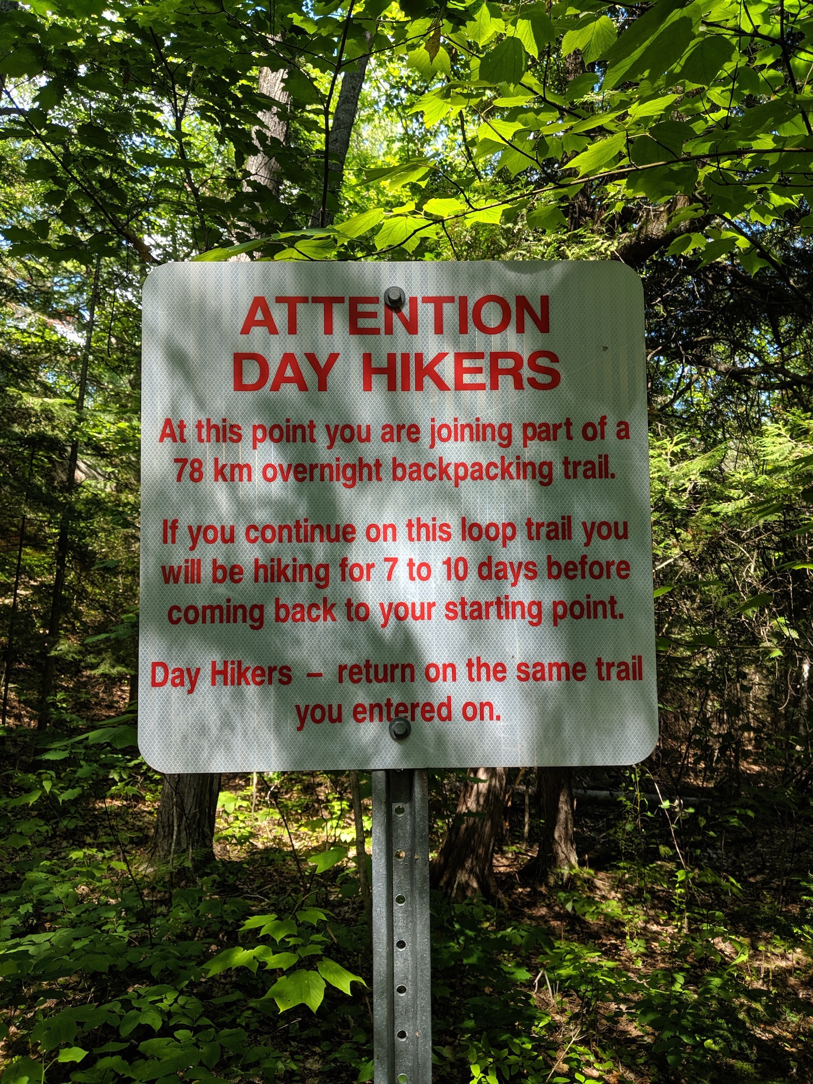 Sign in the woods that reads: "ATTENTION DAY HIKERS: At this point you are joining part of a 78 km overnight backpacking trail. If you continue on this loop trail you will be hiking for 7 to 10 days before coming back to your starting point. Day Hikers -- return on the same trail you entered on." (NB. According to family who work for Ontario Parks, every few years, someone disregards this sign and park staff have to go rescue them.)