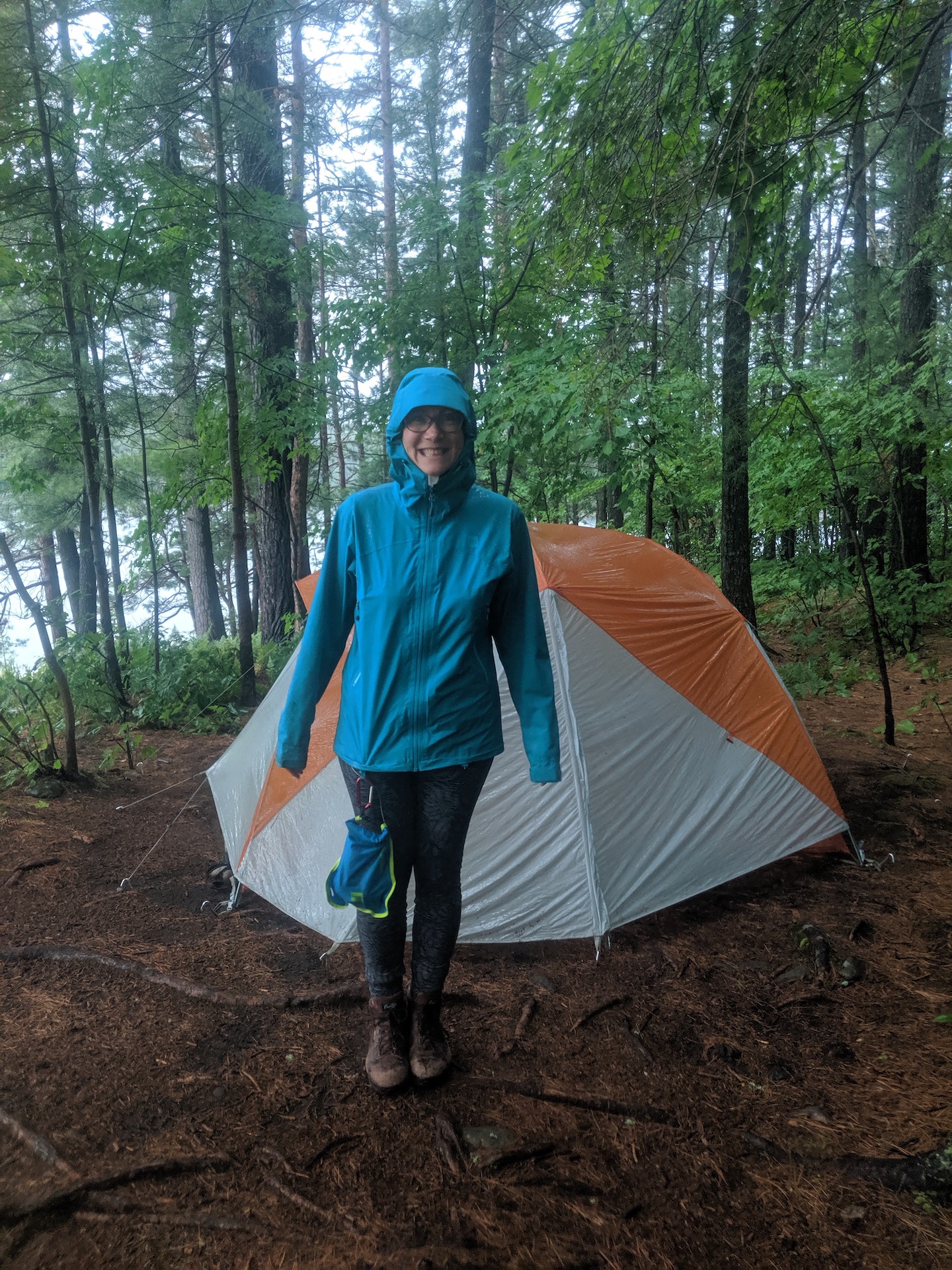 Me in a teal rain jacket and tights with a dry bag clipped to my rain jacket, standing in front of a tent in the woods.