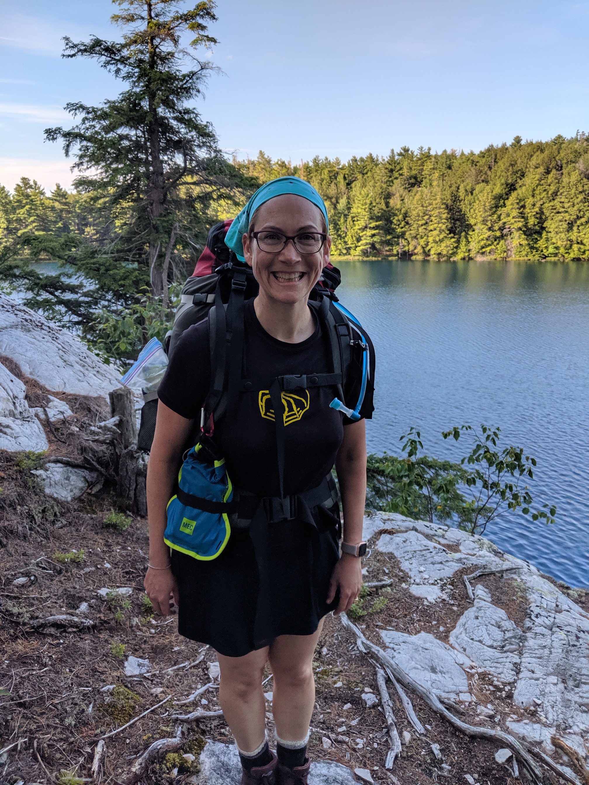 Me in a black Tshirt with a yellow spider on it (an old Torontula jersey from a long-ago phase of my life), a black skort, a teal bandanna, and a hiking pack with a blue dry bag tucked into the waist strap. I am standing in front of a lake surrounded by trees.