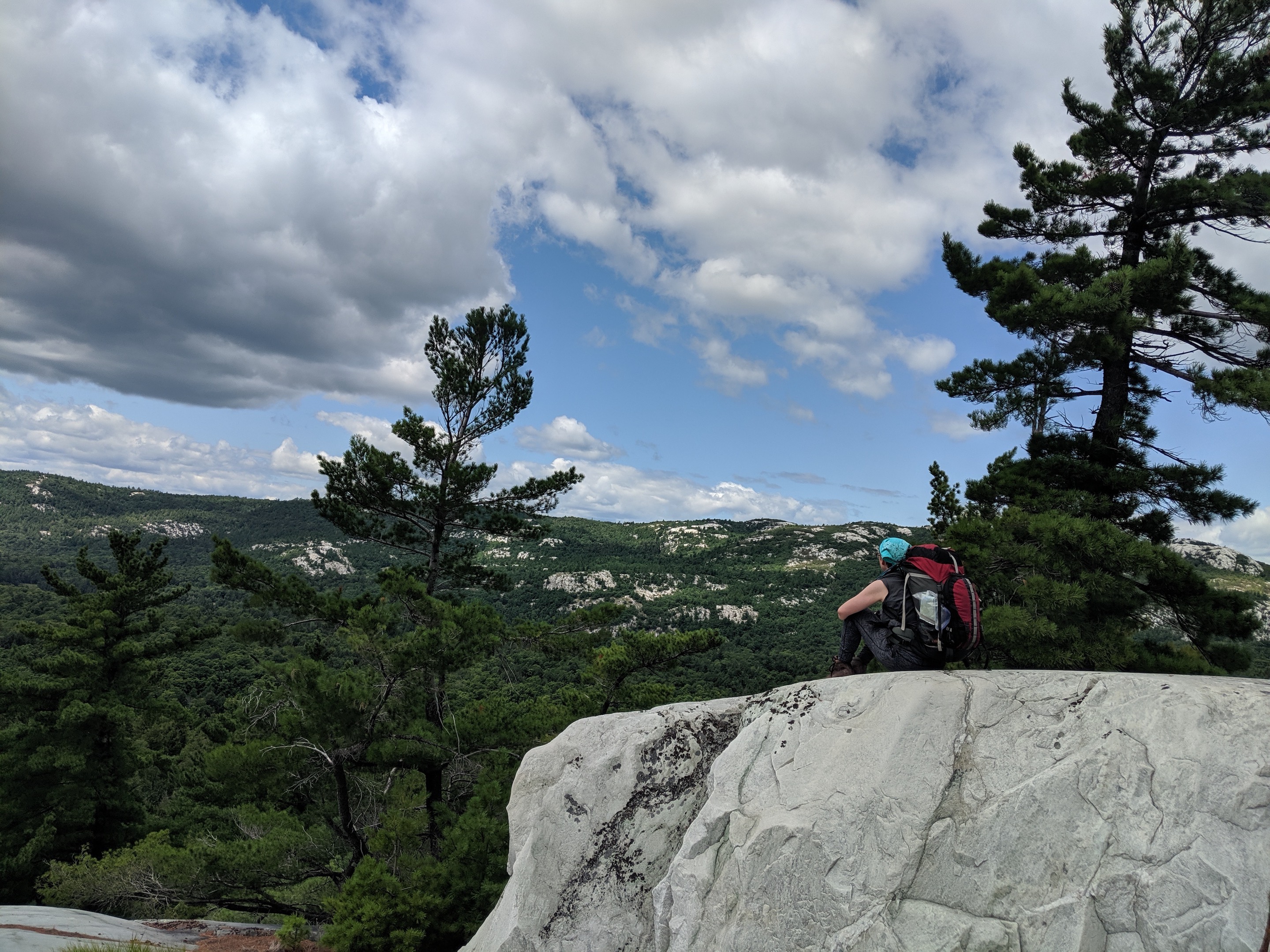 Me sitting on a big white rock with a pack on my back, looking out over trees and blue sky with scattered clouds.