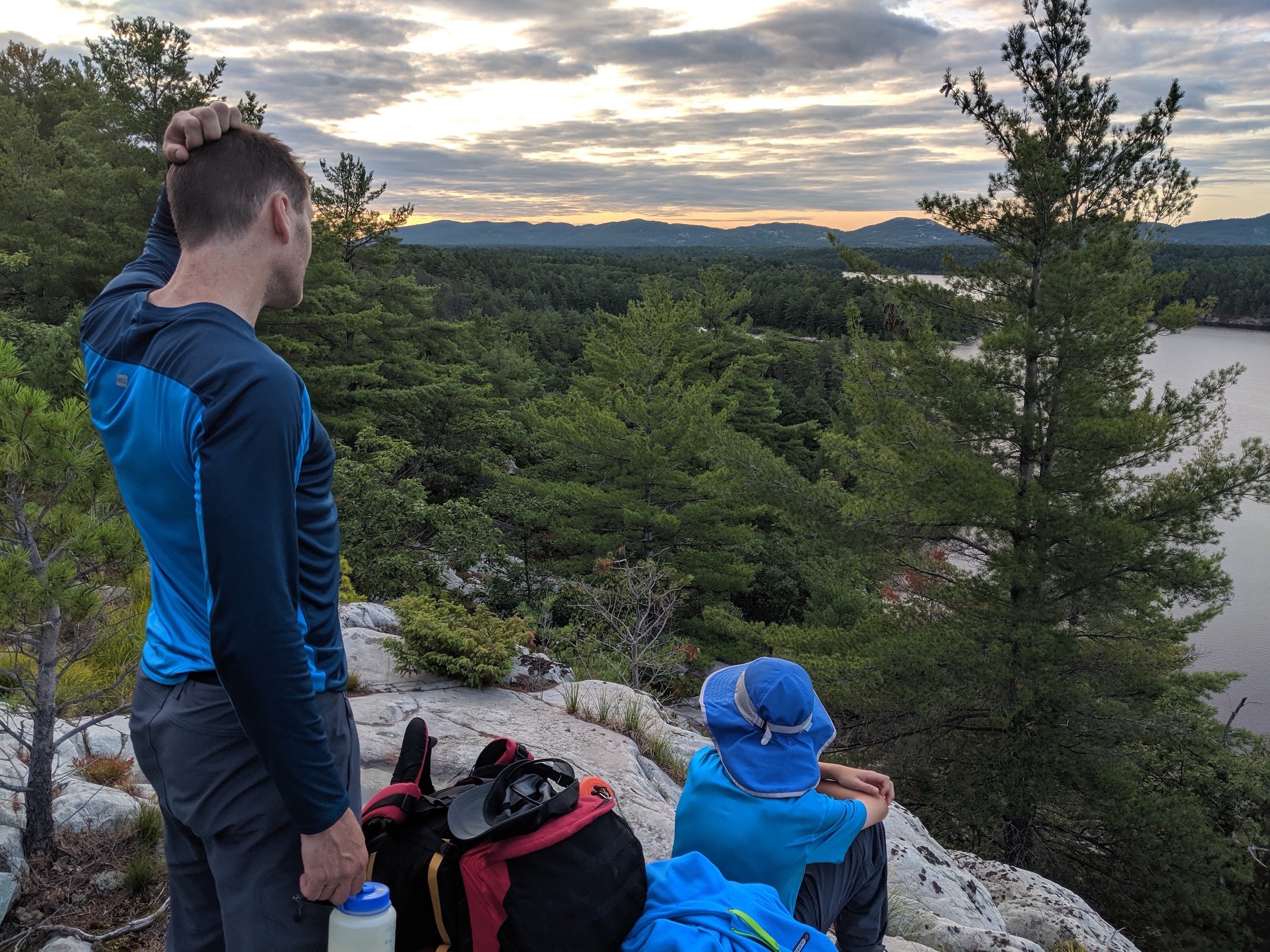 My husband and 13-year old up high on a rock at sunrise, overlooking trees and lakes.