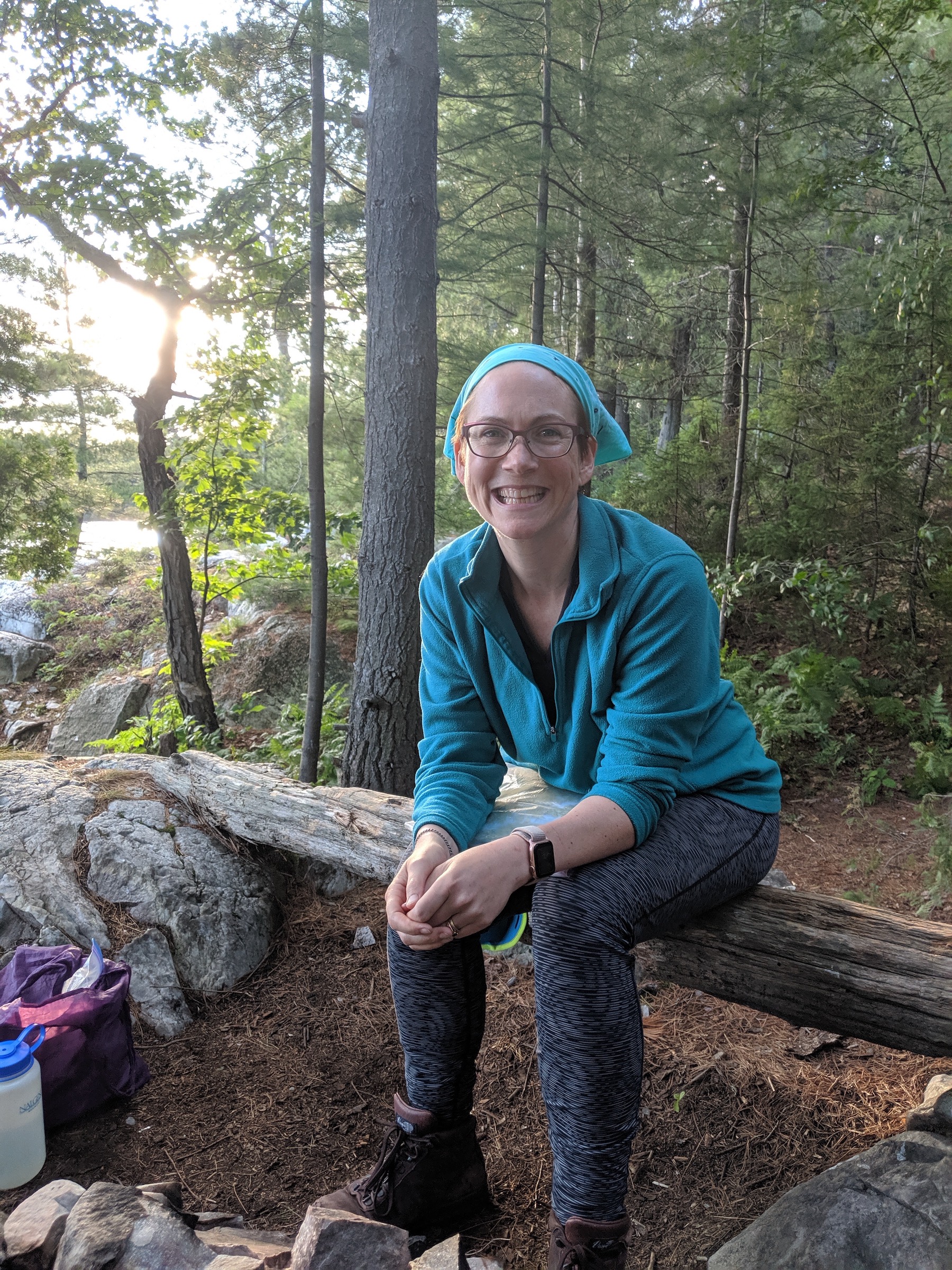 Me sitting on a log in grey/black tights, a teal fleece and bandanna, and a big smile, enjoying being almost done on day 4, feeling OK again and having finally found settings that worked.