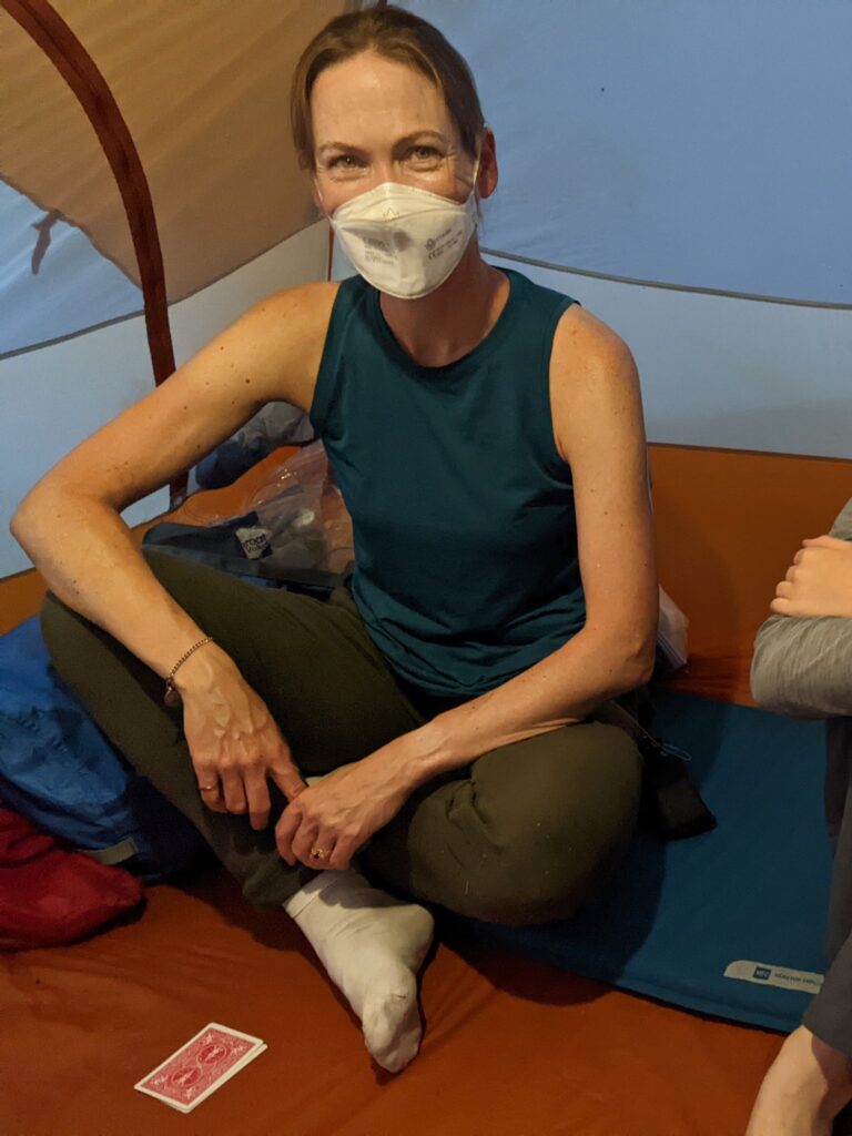 A white woman with brown hair pulled back and a mask on her face sitting cross legged in a tent with cards on the ground in front of her.